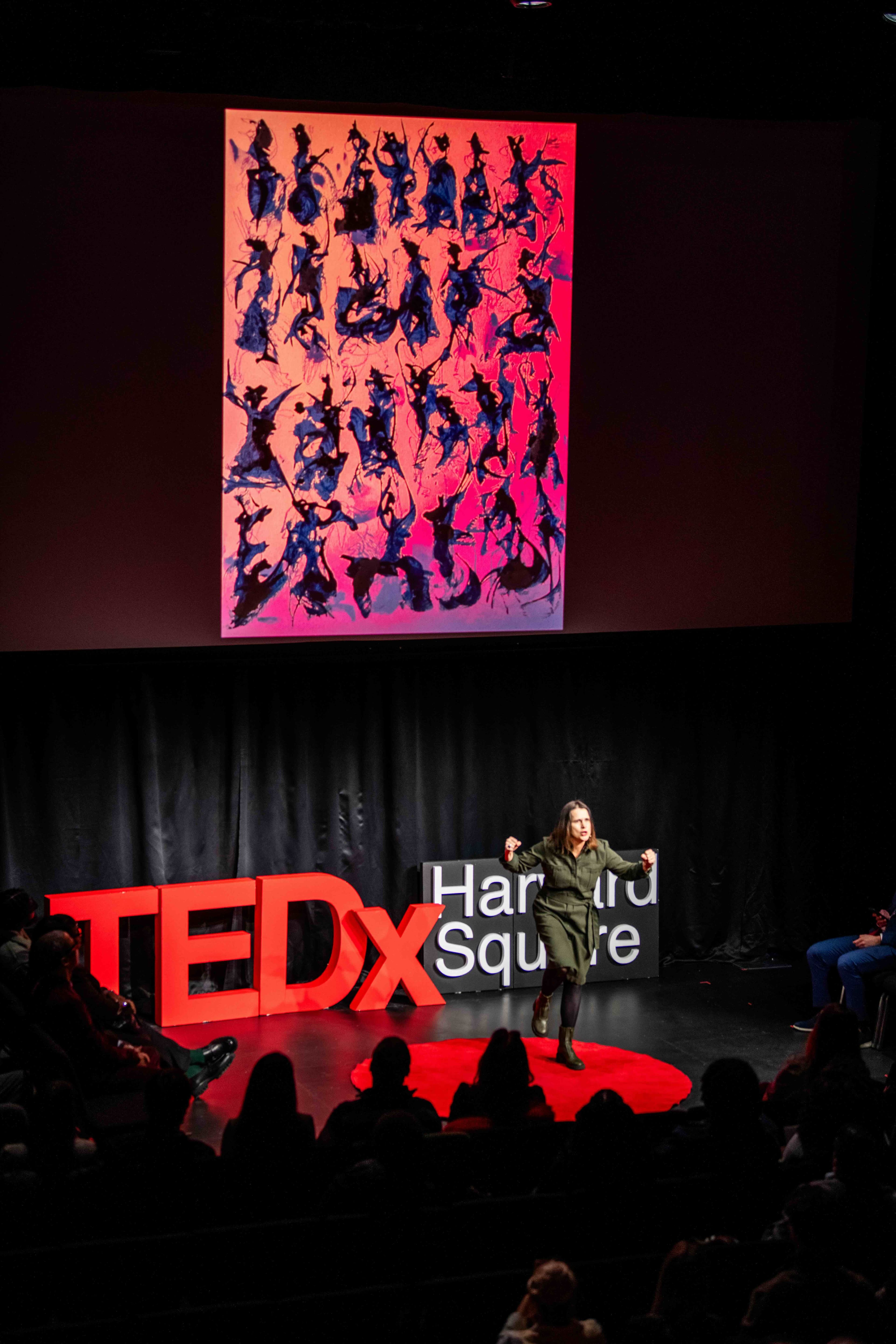 A speaker on the TEDxHarvardSquare stage, beside the red TEDx letters and beneath a large projected artwork, with the audience in the foreground.