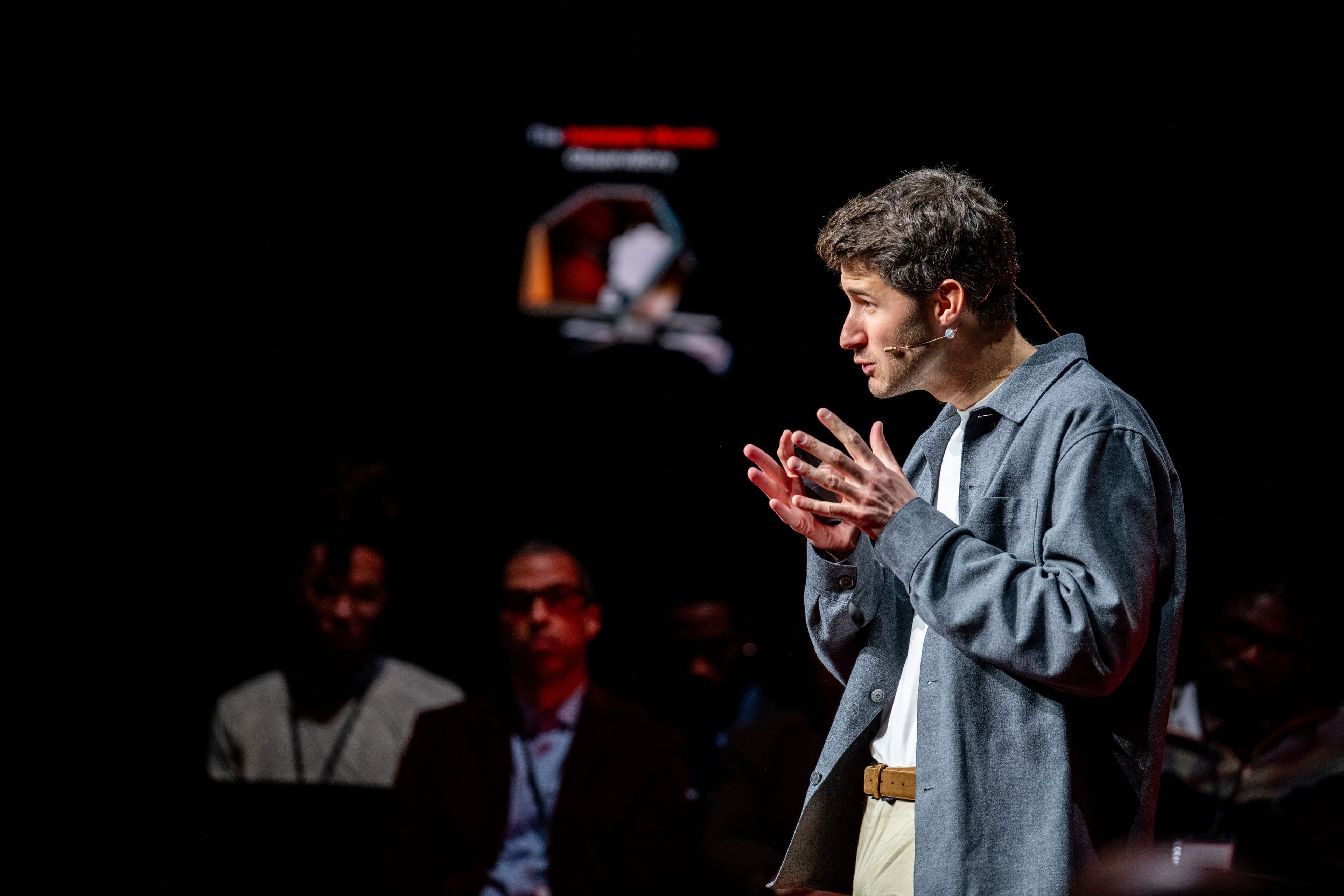 A speaker in a grey overshirt gestures with both hands mid-talk.