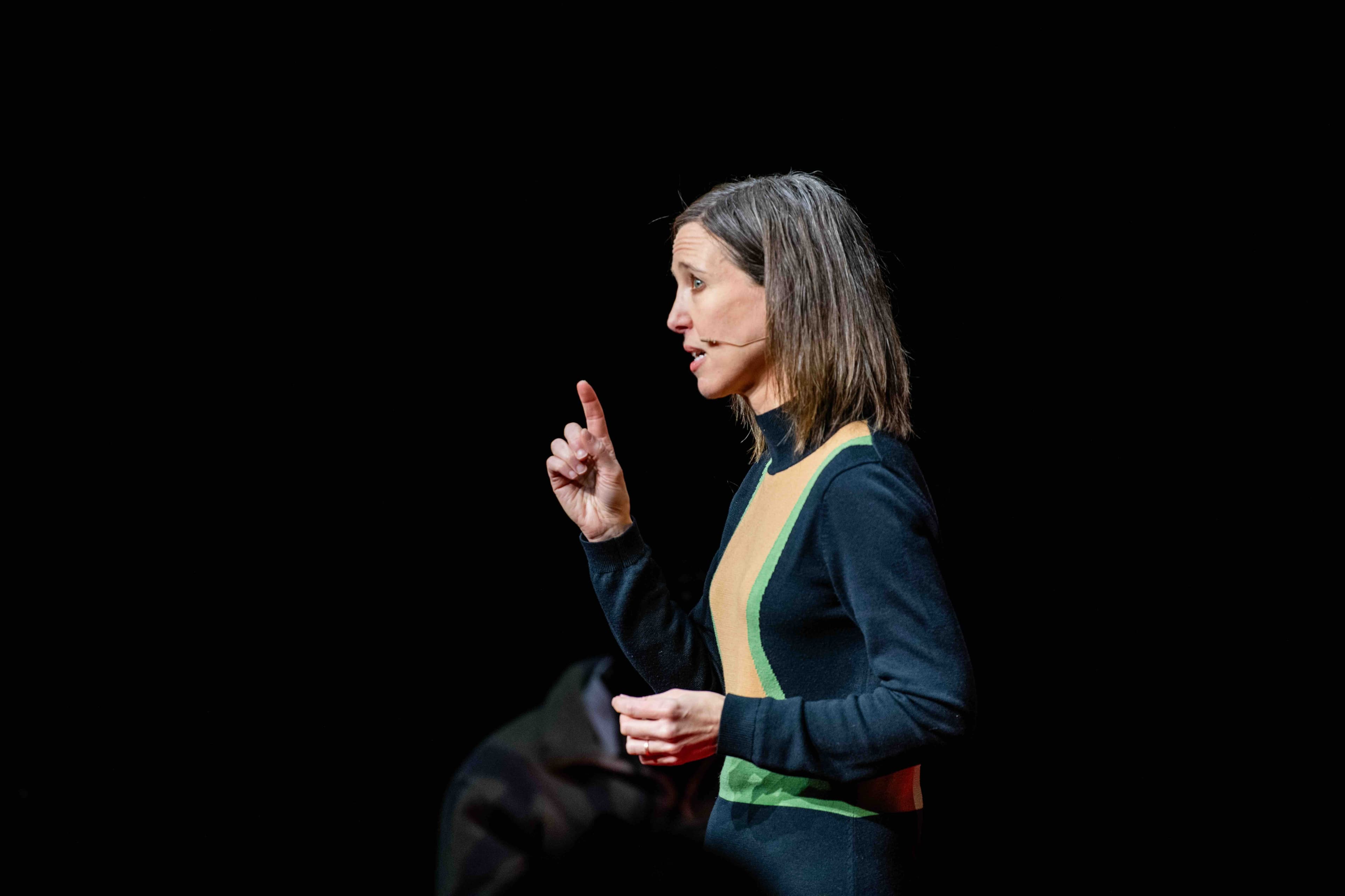 A speaker in profile makes a point with one raised finger against a dark stage.