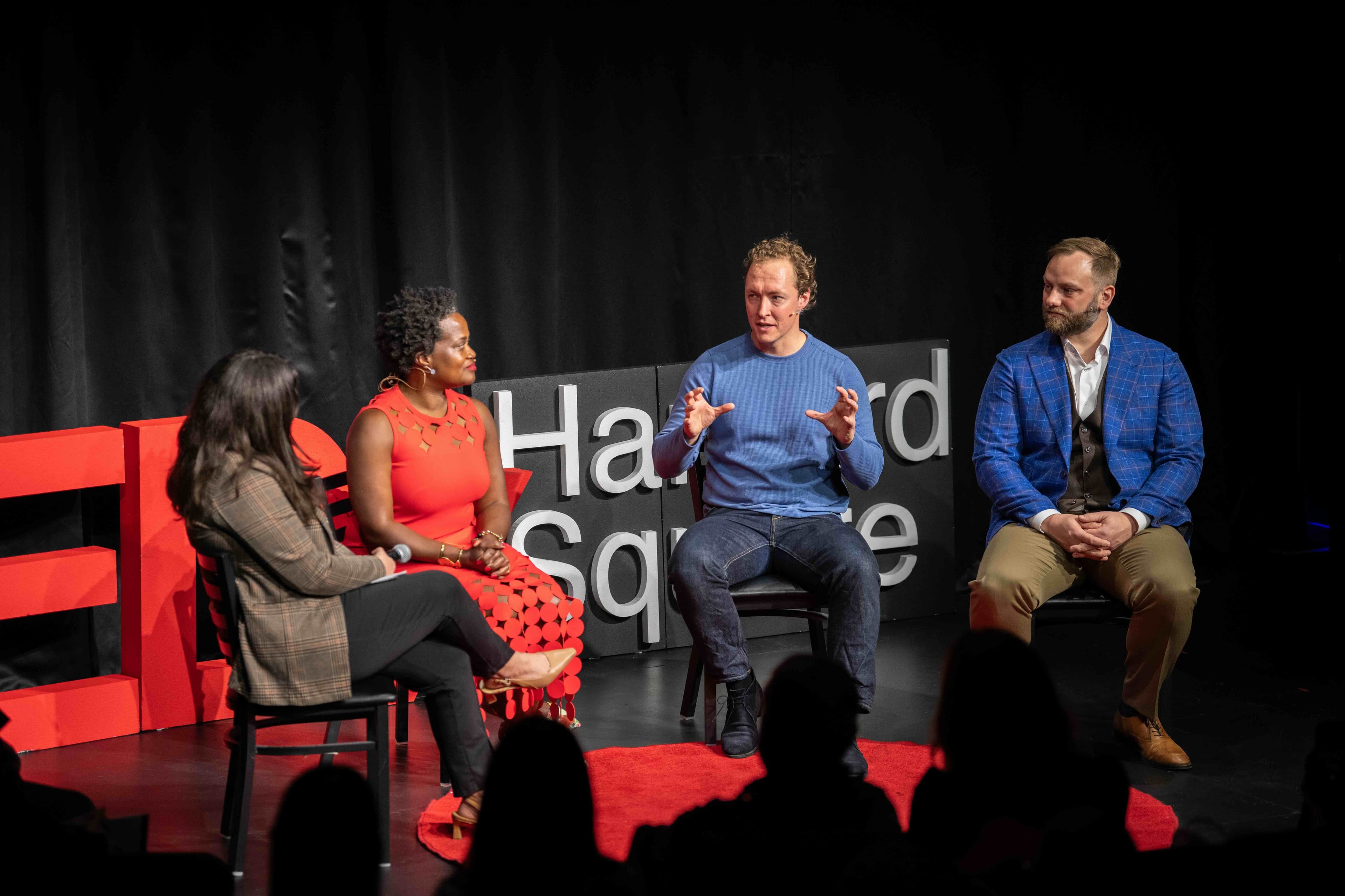 Four panelists in conversation on the TEDxHarvardSquare stage, beside the red TEDx letters.